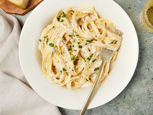 Copycat Olive Garden alfredo in a bowl with a fork, and in the surroundings, a white table napkin, a block of parmesan on a wooden board, and a glass with a drink