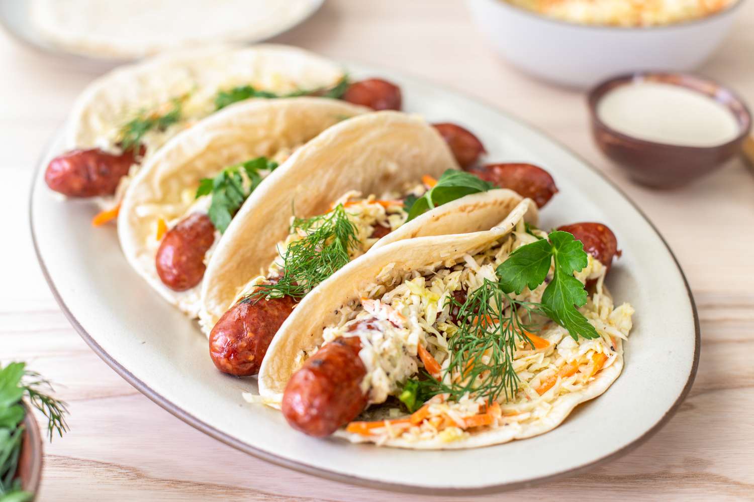 Plate of Ronto Wraps Topped With Herbs, Slaw, and Dressing, and in the Background, a Bowl of Coleslaw, a Plate With Pita Bread, and a Bowl of Dressing