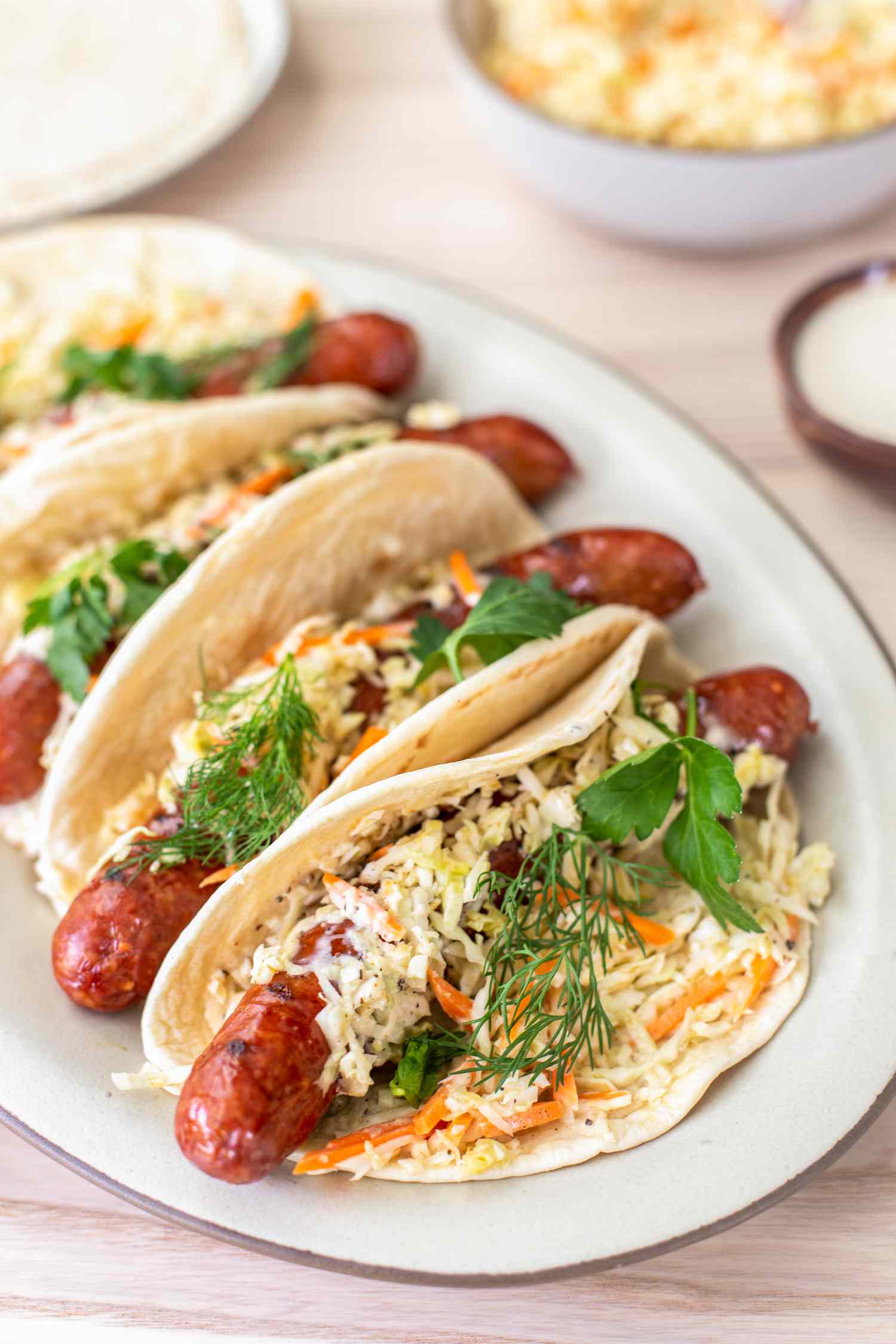 Plate of Ronto Wraps Topped With Herbs, Slaw, and Dressing, and in the Background, a Bowl of Coleslaw, a Plate With Pita Bread, and a Bowl of Dressing