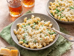 Bowl of Creamy Cavatappi Topped With Shredded Parmesan With a Fork and Sitting on an Olive Kitchen Towel. In the Surroundings, Another Bowl With a Serving, a Small Bowl With Blocks of Parmesan, and a Two Glasses.