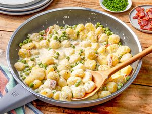 Creamy Skillet Gnocchi With Bacon and Peas in a skillet with a spoon at a table setting with a stack of plates, a bowl of chopped chives, and a small plate of bacon pieces