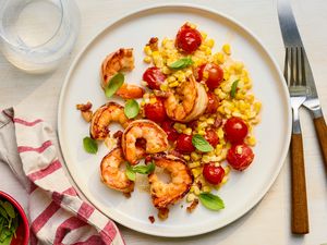 Plate of creamy skillet shrimp with corn, pancetta, and tomatoes at a table setting with a glass of water, a white and red table napkin on the counter, and a bowl of basil