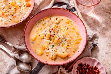 Bowl of creamy vegetable dumpling soup, and in surroundings, two spoons on the counter, another serving in a bowl, a small bowl of crushed peppers, a bowl of shredded parmesan, and a grey and black kitchen linen