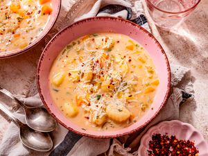 Bowl of creamy vegetable dumpling soup, and in surroundings, two spoons on the counter, another serving in a bowl, a small bowl of crushed peppers, a bowl of shredded parmesan, and a grey and black kitchen linen