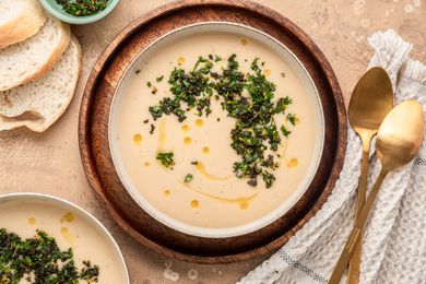 Two Bowls of Creamy White Bean Soup with Sage Gremolata, Surrounded by Slices of Bread on the Counter, a Bowl of Sage Gremolata, and Spoons on a Kitchen Towel