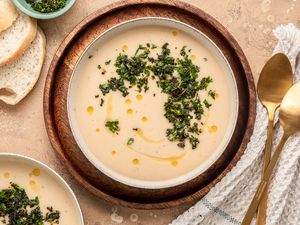 Two Bowls of Creamy White Bean Soup with Sage Gremolata, Surrounded by Slices of Bread on the Counter, a Bowl of Sage Gremolata, and Spoons on a Kitchen Towel