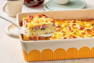 Slice of Denver omelette hashbrown casserole lifted from a casserole dish, and in the surroundings, a stack of plates and a mug