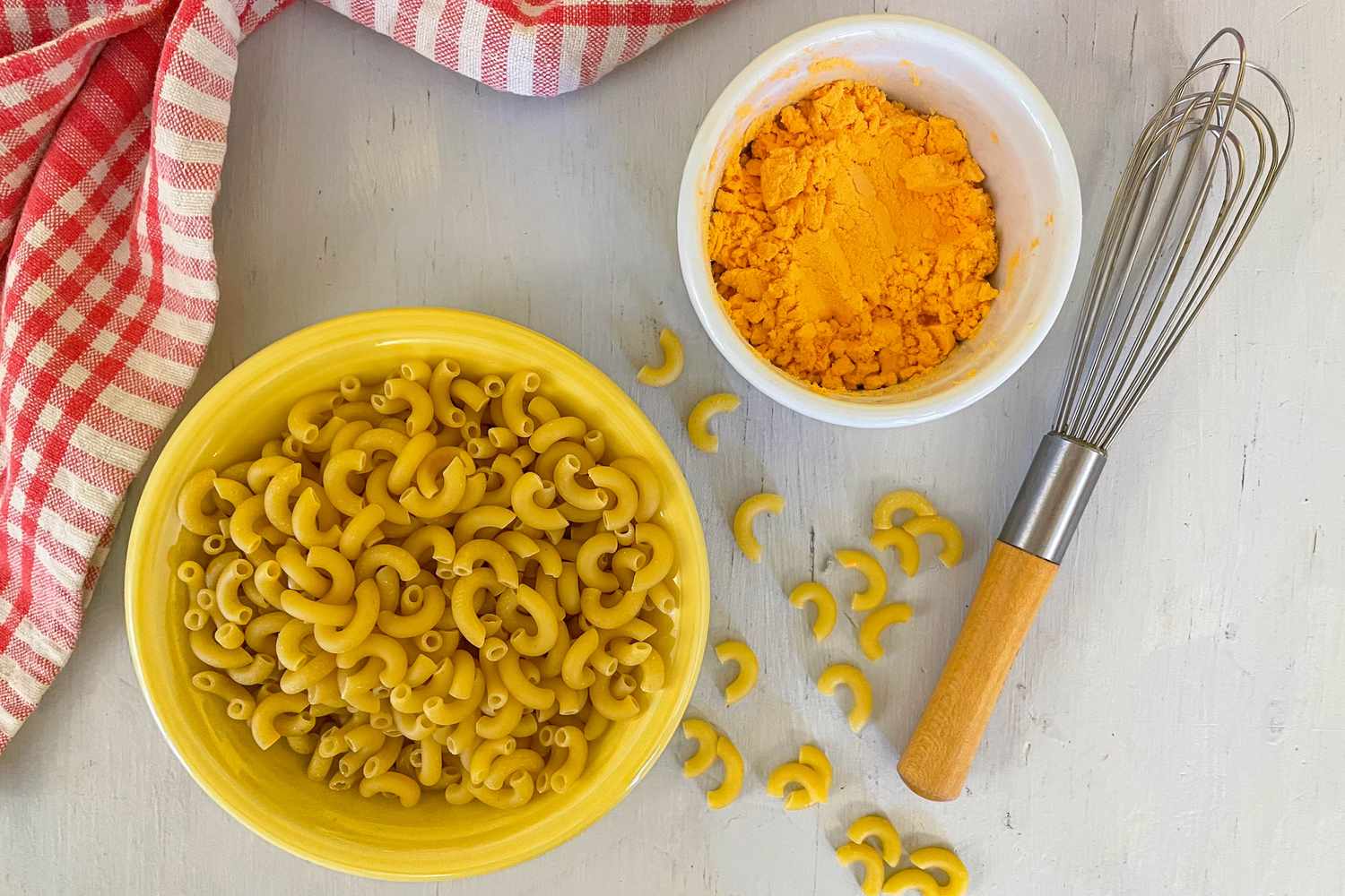 Bowl of Uncooked Macaroni With Some Rogue Pieces Scattered on the Counter, a Bowl of Dry Cheese Powder, a Whisk, and a Red and White Checkered Kitchen Towel