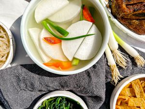 Bowl of Dongchimi Surrounded by Bowls of Banchan (Side Dishes) and a Bowl of Korean Short Ribs. On the Table, a Table Napkin and Some Whole Green Onions.