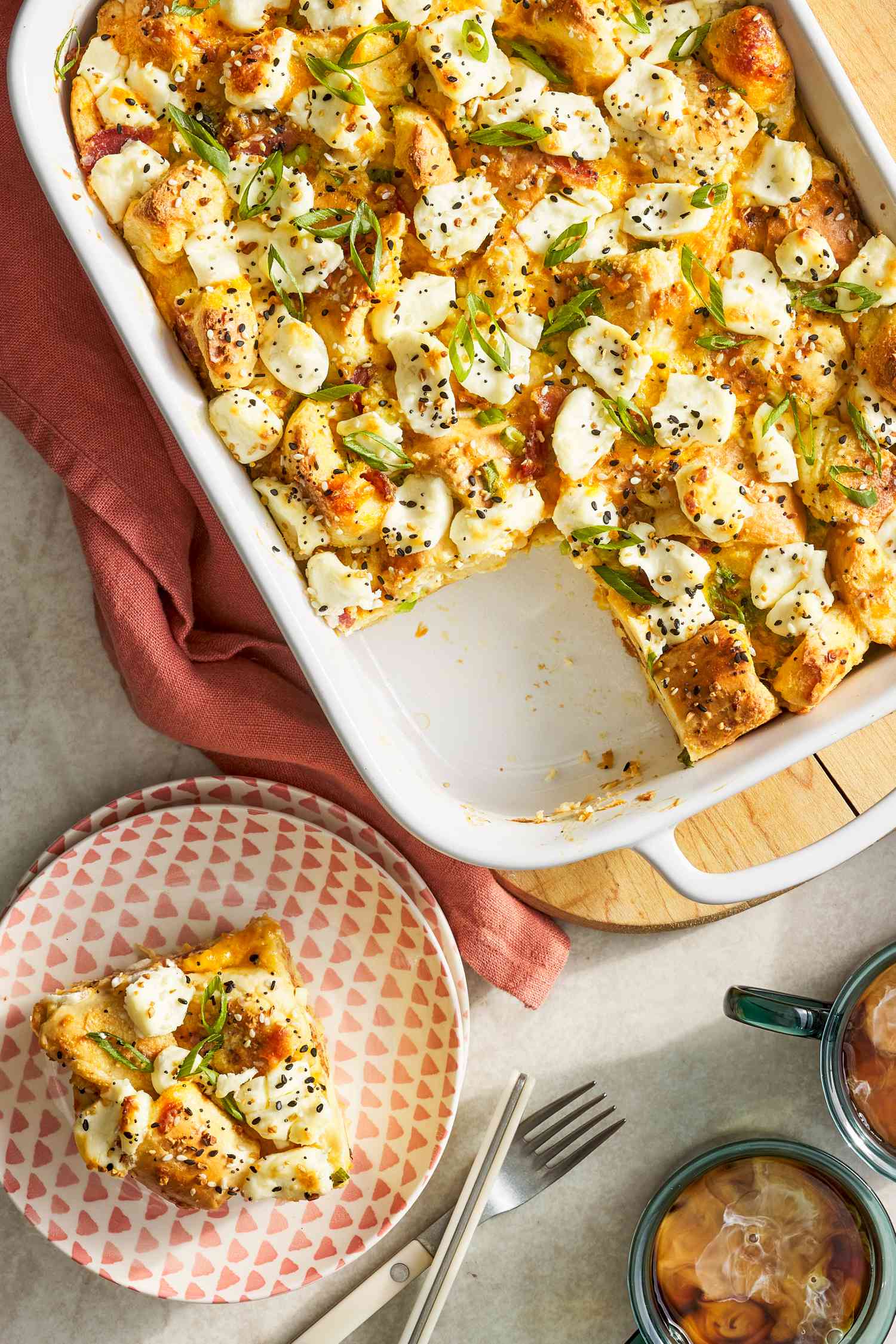 Everything bagel sandwich casserole in a casserole dish at a table setting with a serving on a plate, utensils, and cups of coffee