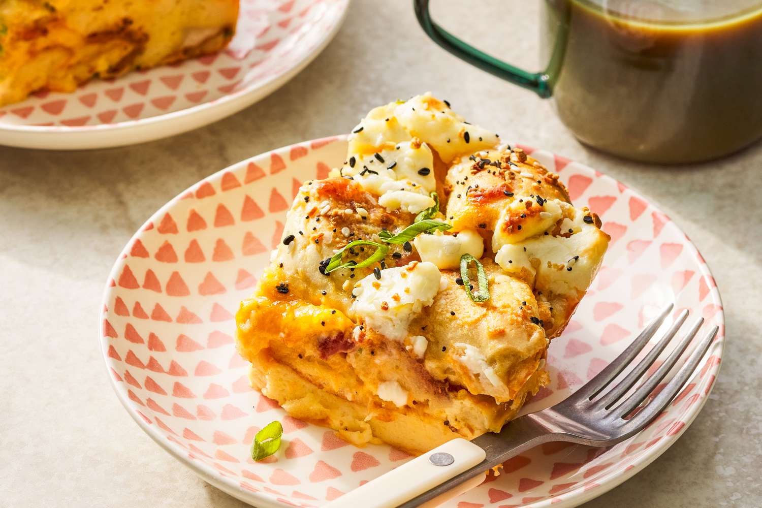 Serving of everything bagel sandwich casserole on a small plate with a fork next to a cup of coffee and another serving on a plate 