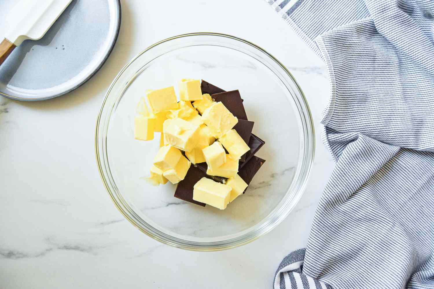 Bowl of chocolate and butter pieces, and next to the bowl, a spatula resting on a small plate and a blue and white kitchen towel for fluffernutter brownie recipe