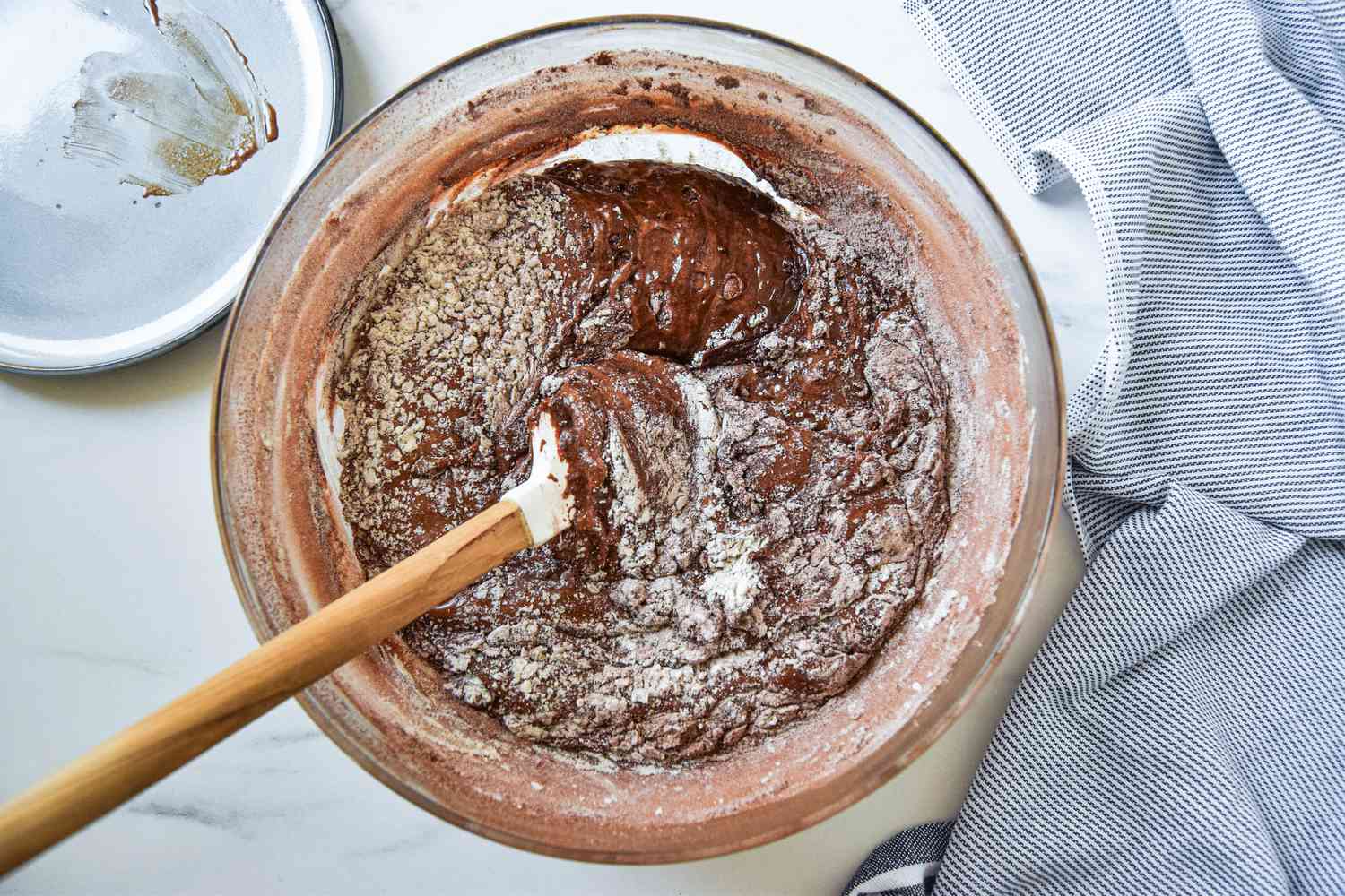 Dry ingredients carefully folded into the wet ingredients using a spatula, all next to a small plate and a blue and white striped kitchen towel for fluffernutter brownies recipe