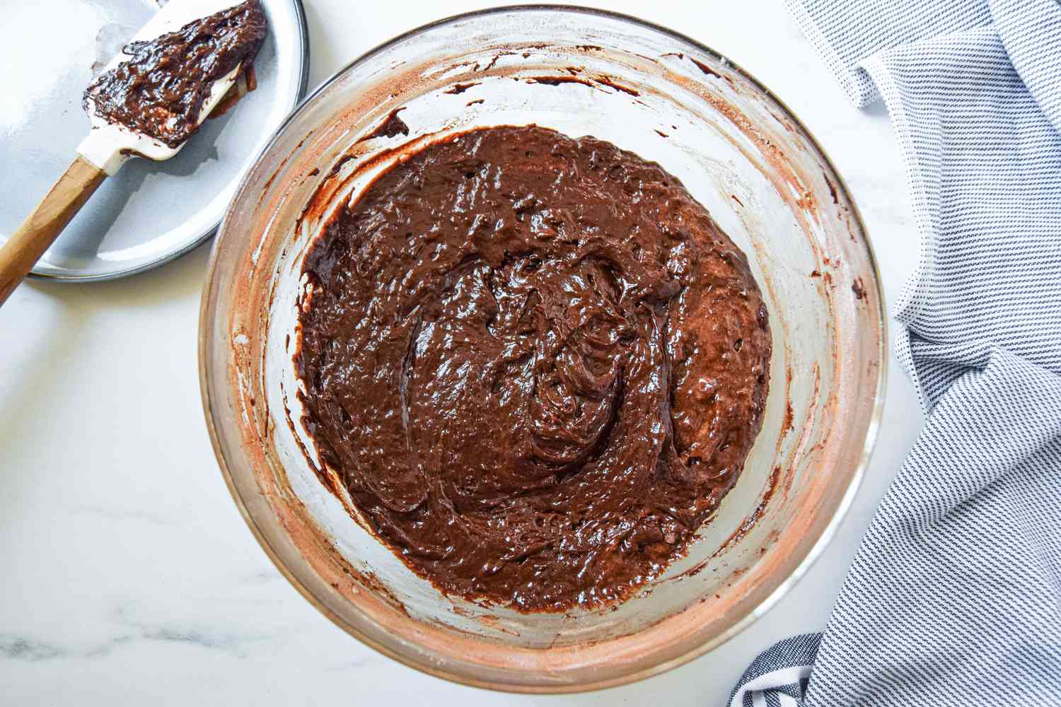 Fluffernutter brownie batter in a bowl next to a small plate with the spatula and a blue and white striped kitchen towel