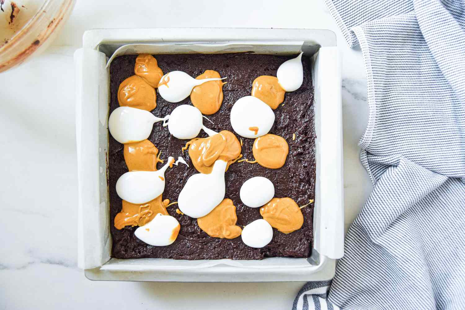 Small dollops of peanut butter and marshmallow fluff added to fluffernutter brownie butter in the baking pan, and next to it, the blue and white striped kitchen towel