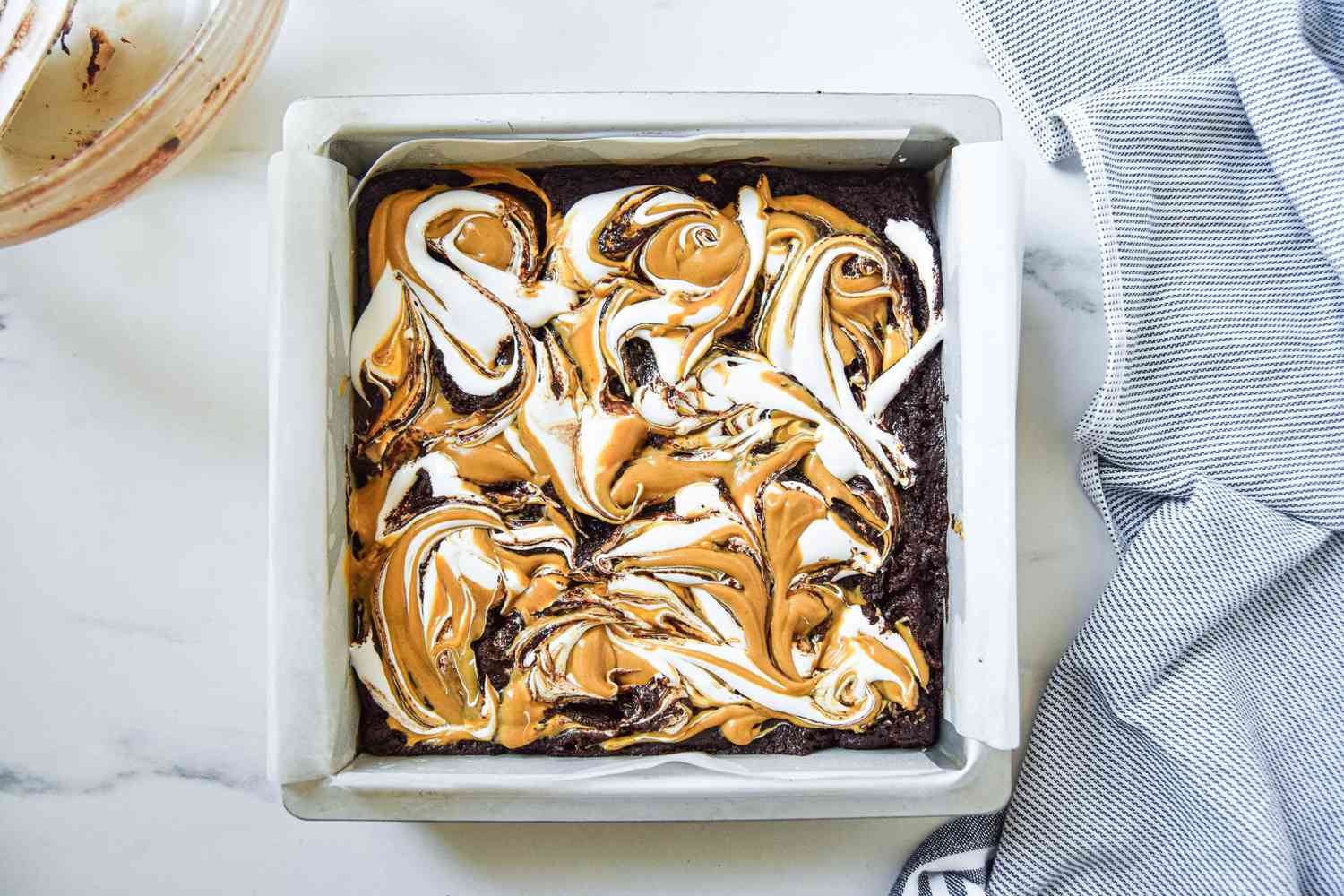 Peanut butter and marshmallow fluff swirled into fluffernutter brownie butter in the baking pan, and next to it, the blue and white striped kitchen towel