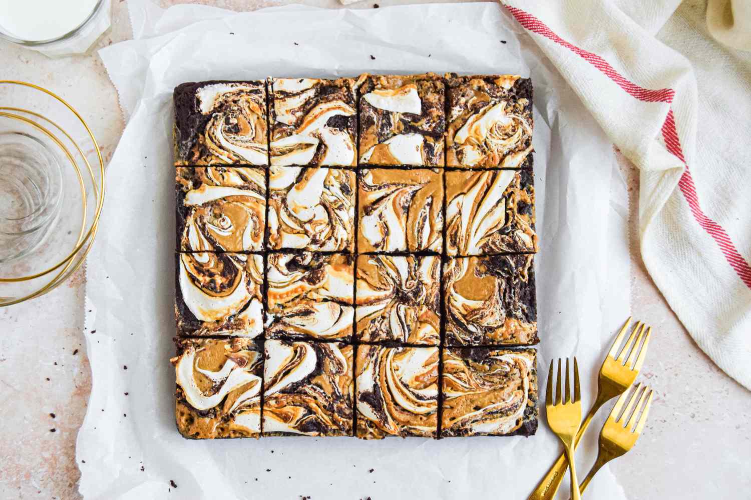 Fluffernutter brownie cut into 16 slices on a piece of parchment paper, and in the surroundings, a white kitchen towel, forks on the counter, a stack of small plates, and a glass of milk