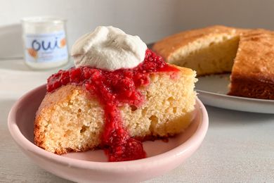 Slice of French yogurt cake with raspberry sauce and a dollop of whipped cream on a plate next to a cake pan with more and a bottle of Oui yogurt