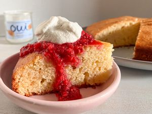 Slice of French yogurt cake with raspberry sauce and a dollop of whipped cream on a plate next to a cake pan with more and a bottle of Oui yogurt