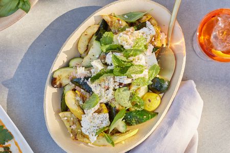 Bowl of Grilled Zucchini and Yellow Squash with Champagne Vinaigrette with a Spoon Surrounded by a Drink on a Table 