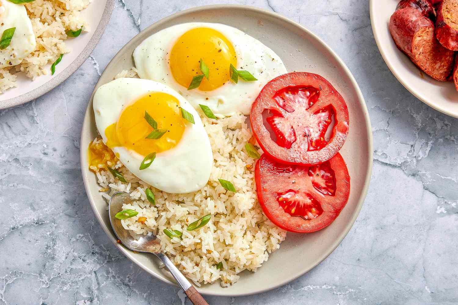 Plate of Garlic Fried Rice (Sinangag na Kanin) with sunny side eggs and sliced tomatoes, and in the surroundings, another plate with another serving of rice and eggs and a plate of longanisa (Filipino sausage)