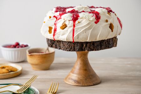 Gingersnap Pavlova with Cranberry Sauce Sitting on a Wooden Cake Stand, Surrounded by a Bowl of More Sauce, a Bowl with Cranberries, and a Plate with Gingersnaps