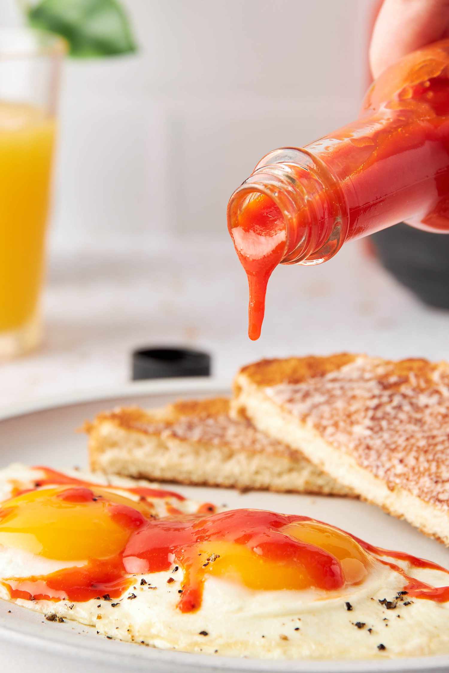 Homemade Hot Sauce Poured From a Bottle Onto Sunny Side Eggs With a Side of Toast, and in the Background, a Glass of Orange Juice and a Kitchen Plant