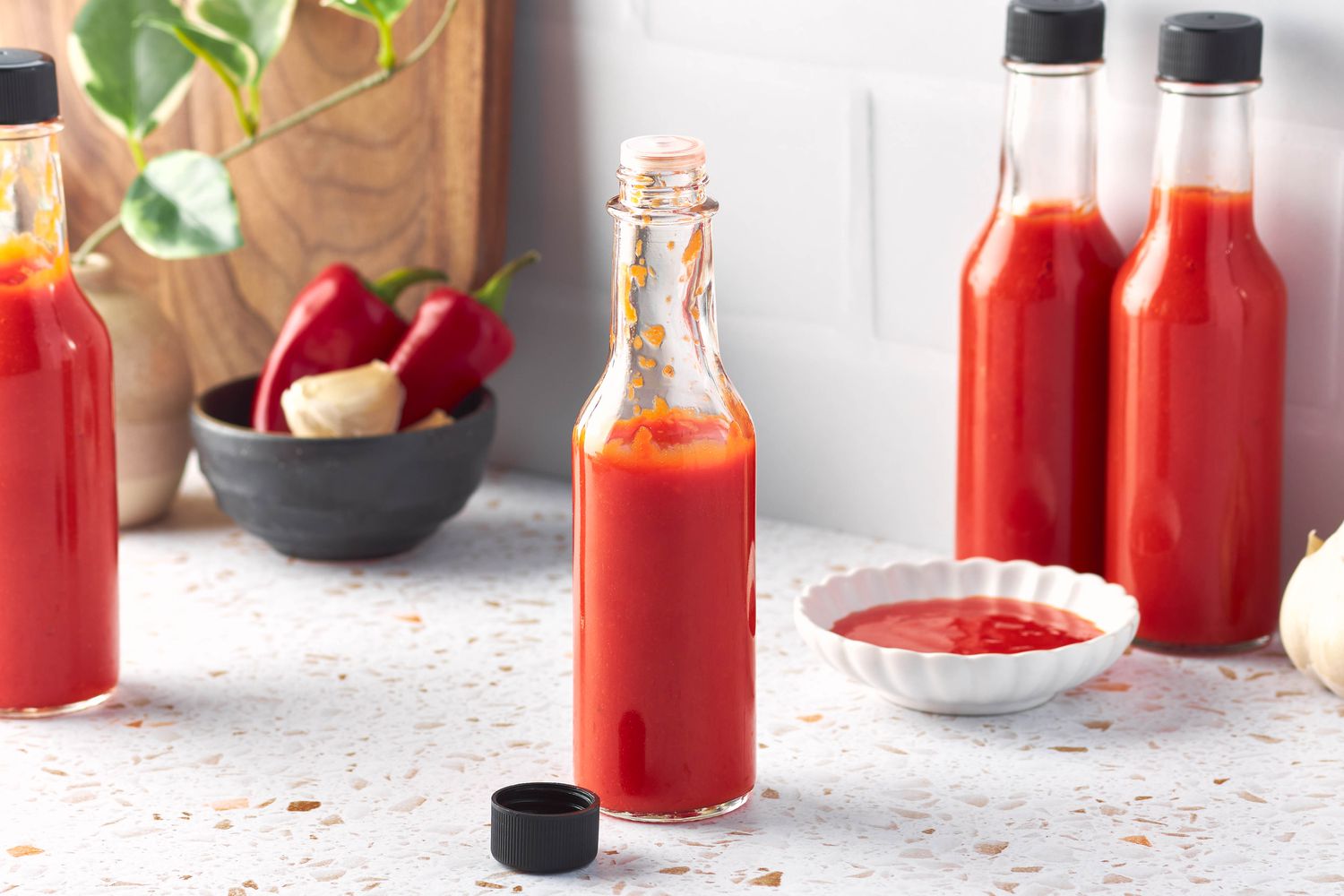 Bottle of Homemade Hot Sauce Open With the Cap Next to the Bottle on the Counter, and in the Surroundings, a Small Bowl of Hot Sauce, More Bottles of Homemade Hot Sauce, a Bowl of Red Fresno and Fresh, Whole Garlic Cloves, and a Kitchen Plant