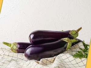 Three Eggplants Stacked Together on a White and Blue Checkered Kitchen Towel Next to Some Parsley. Edges of Photo Decorated With Yellow Polka Dotted Illustrations