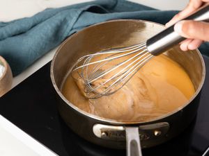 Roux whisked in a saucepan on an induction stove, and in the surroundings, a bowl of flour and a blue kitchen towel