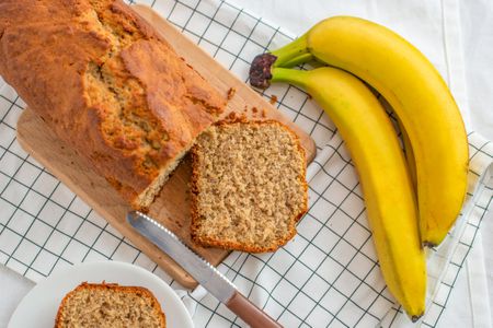 Banana bread loaf with a slice cut laying on the cutting board next to some bananas