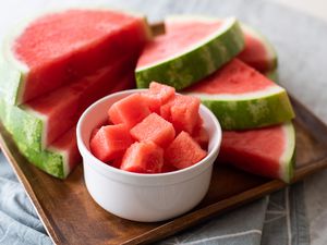 Bowl of watermelon cubes with watermelon slices behind it on a wood serving platter