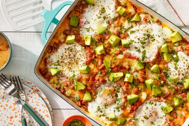 Huevos rancheros casserole at a table setting with a stack of plates and utensils, a cup of coffee, a small bowl of herbs, and a spatula