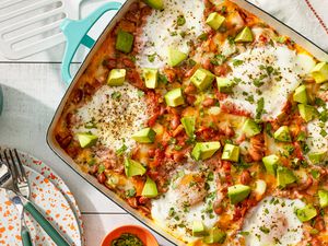 Huevos rancheros casserole at a table setting with a stack of plates and utensils, a cup of coffee, a small bowl of herbs, and a spatula