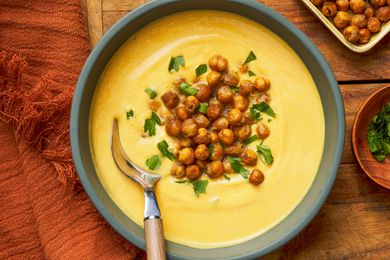 Bowls of hummus next to a small bowl of roasted chickpeas, another small bowl with chopped parsley, and a burnt orange kitchen towel, all on a wooden table