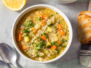 Bowl of rice and vegetable soup, and in the surroundings, slices of toast on a white and blue kitchen towel, a halved lemon, and utensils on the counter