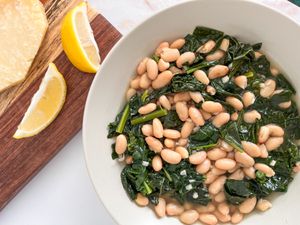 Bowl of Italian beans and greens next to a pot with more, a wooden board with lemon wedges, and a block of parmesan