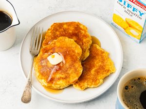 Jiffy Pancakes topped with syrup and a slab of butter on a plate at a table setting wtih a container of maple syrup, a box of jiffy muffin mix, and a mug of coffee