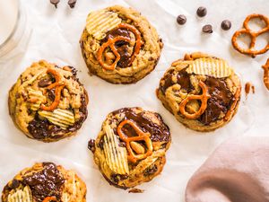 Kitchen Sink Cookies, Pretzels, Chocolate Chips, and a Glass of Milk on a Piece of Crumpled Parchment Paper and a Mauve Kitchen Linen