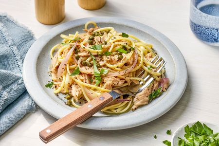Linguine with tuna and capers on a plate at a table setting with a bowl of parsley, a denim table napkin, salt and pepper shakers, and a glass of water