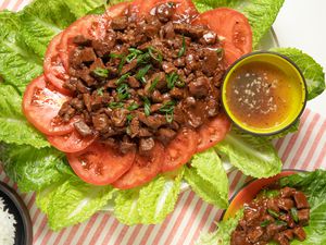 Platter of Lok Lak Served Over Sliced Tomatoes and Lettuce at a Table Setting With a Bowl of Rice, a Plate With a Serving of Lok Lak, and a Bowl of Pepper-lime Sauce 