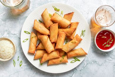 Plate of Lumpia Cut in Half and Topped With Sliced Green Onions, and in the Surroundings, a Bowl of Rice, a Bowl of Sweet and Sour Sauce With Green Onions, and Two Glasses of a Fizzy Drink