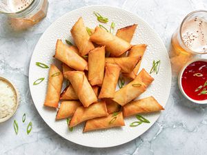 Plate of Lumpia Cut in Half and Topped With Sliced Green Onions, and in the Surroundings, a Bowl of Rice, a Bowl of Sweet and Sour Sauce With Green Onions, and Two Glasses of a Fizzy Drink