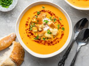 Bowls of Mercimek Corbasi (Turkish Lentil Soup) Topped with Greek Yogurt, Croutons, Pul Bibir Butter, and Parsley, and Next to It, a Small Bowl of Parsley, Slices of Bread, and Two Spoons