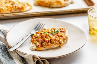 A million dollar chicken breast on a plate with a fork, and in the surroundings, a parchment paper lined baking tray with more, a glass of water, and a table napkin