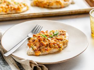 A million dollar chicken breast on a plate with a fork, and in the surroundings, a parchment paper lined baking tray with more, a glass of water, and a table napkin