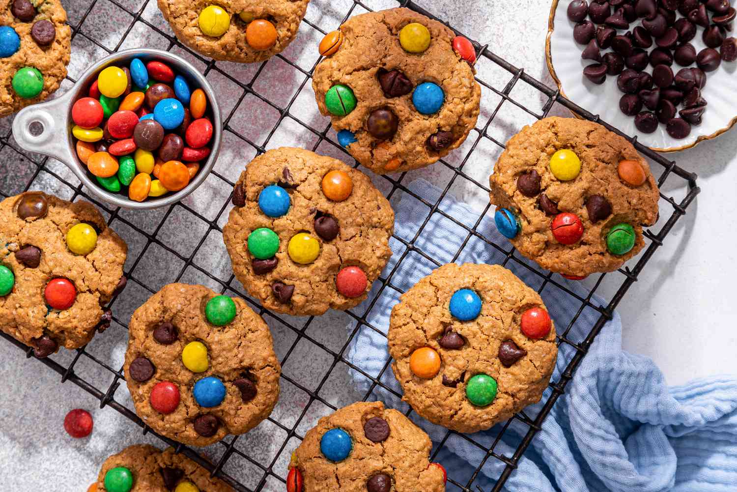 Close-up: monster cookies on a cooling rack with a bowl of M&M's, and on the counter next to the rack, a monster cookie, a bowl of chocolate chips, some rogue M&M's on the counter, and a baby blue kitchen towel