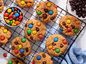 Close-up: monster cookies on a cooling rack with a bowl of M&M's, and on the counter next to the rack, a monster cookie, a bowl of chocolate chips, some rogue M&M's on the counter, and a baby blue kitchen towel