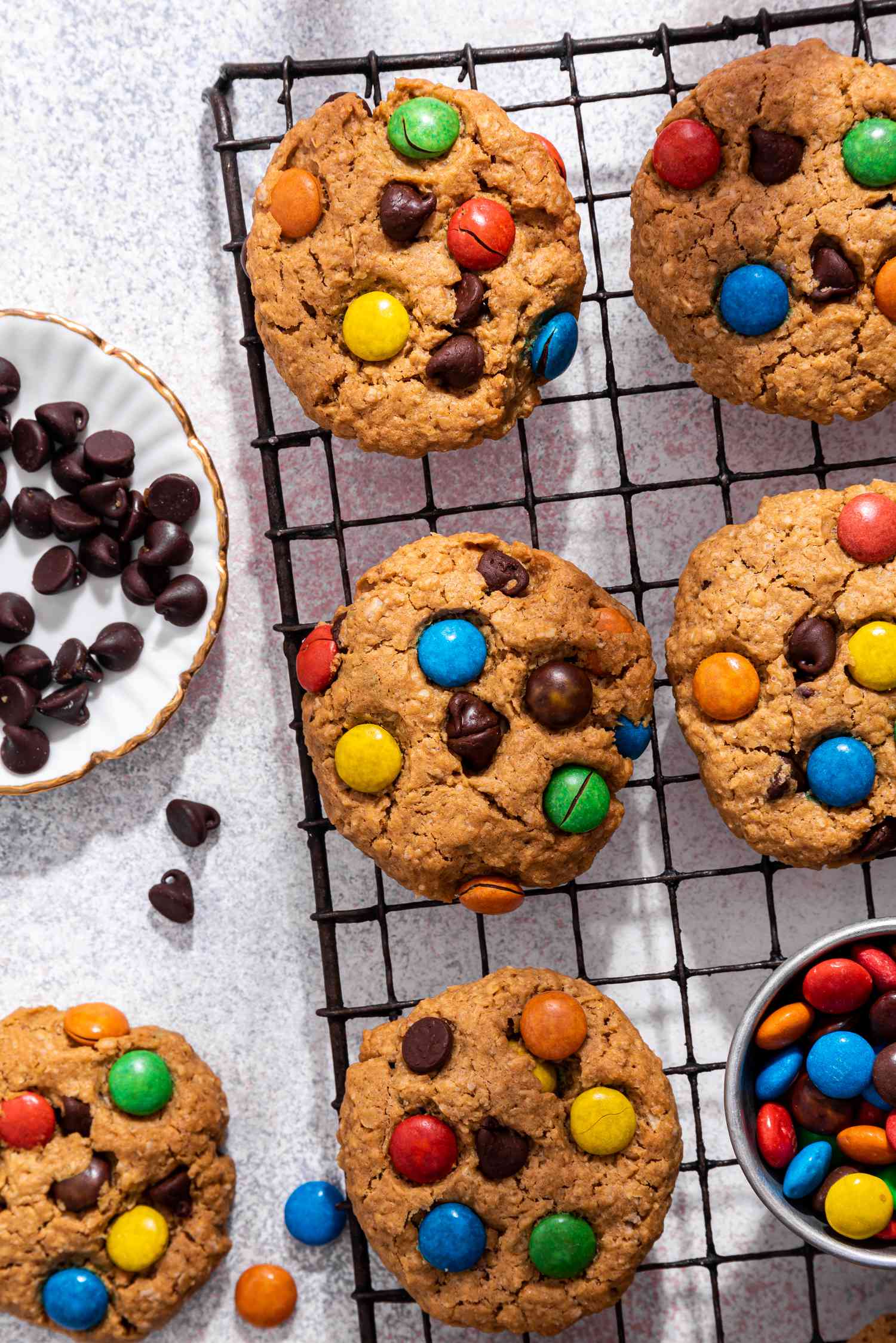 Close-up: monster cookies on a cooling rack with a bowl of M&M's, and on the counter next to the rack, a monster cookie, and a bowl of chocolate chips with a few on the counter