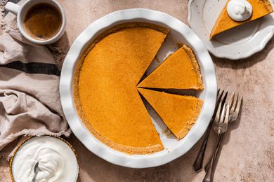 Overhead shot of no-bake pumpkin pie in a pie dish at a rustic setting with a cup of coffe, a grey-ish beige table linen, bowl of whipped cream, and a plate with a slice of pumpkin pie with a dollop of whipped cream